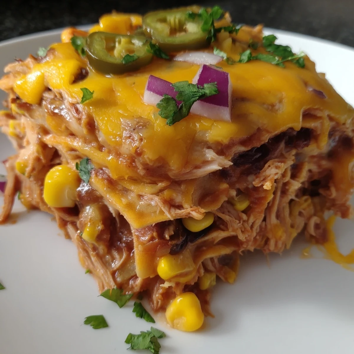 A close-up of layered Chicken Enchilada Casserole with Cheese reveals tender shredded chicken, red sauce, and melted cheese between soft corn tortillas, served with a dollop of sour cream.