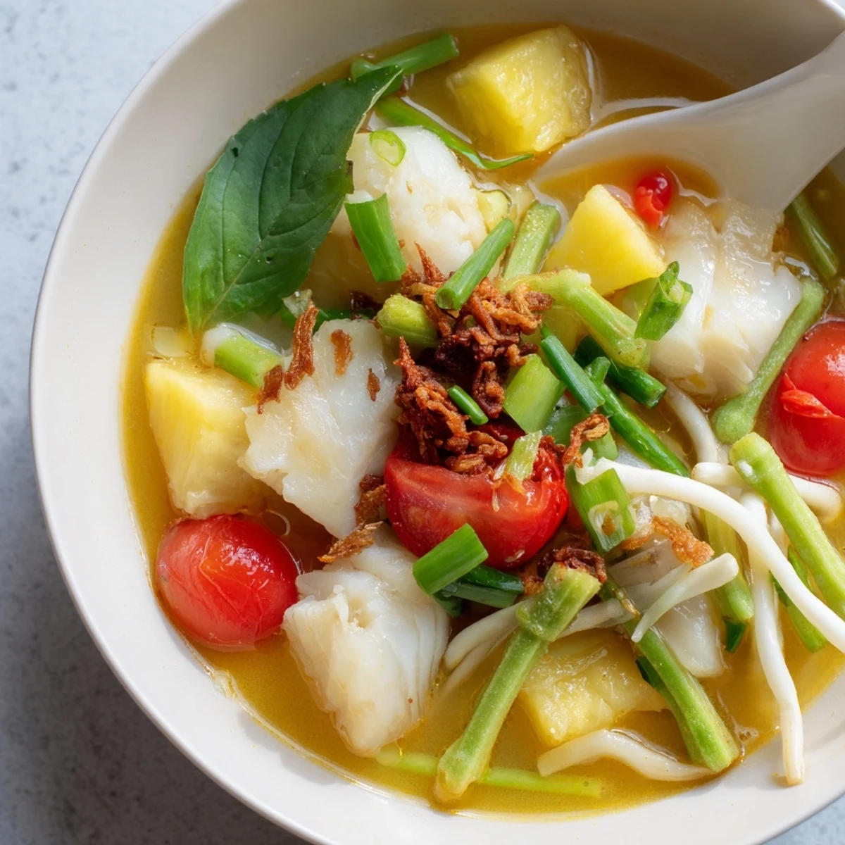 A bowl of Modern Canh Chua sour soup with tender white fish, pineapple chunks, and tomatoes, garnished with fresh herbs.
