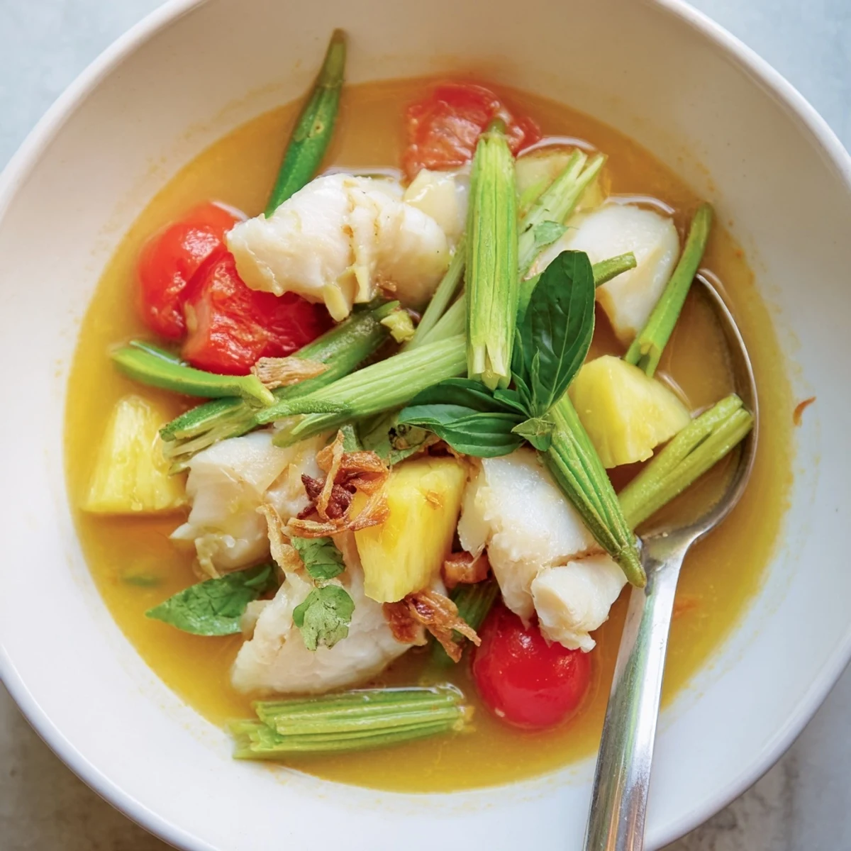 Steaming bowl of Modern Canh Chua topped with Thai basil, cilantro, and fried shallots, served with a lime wedge.